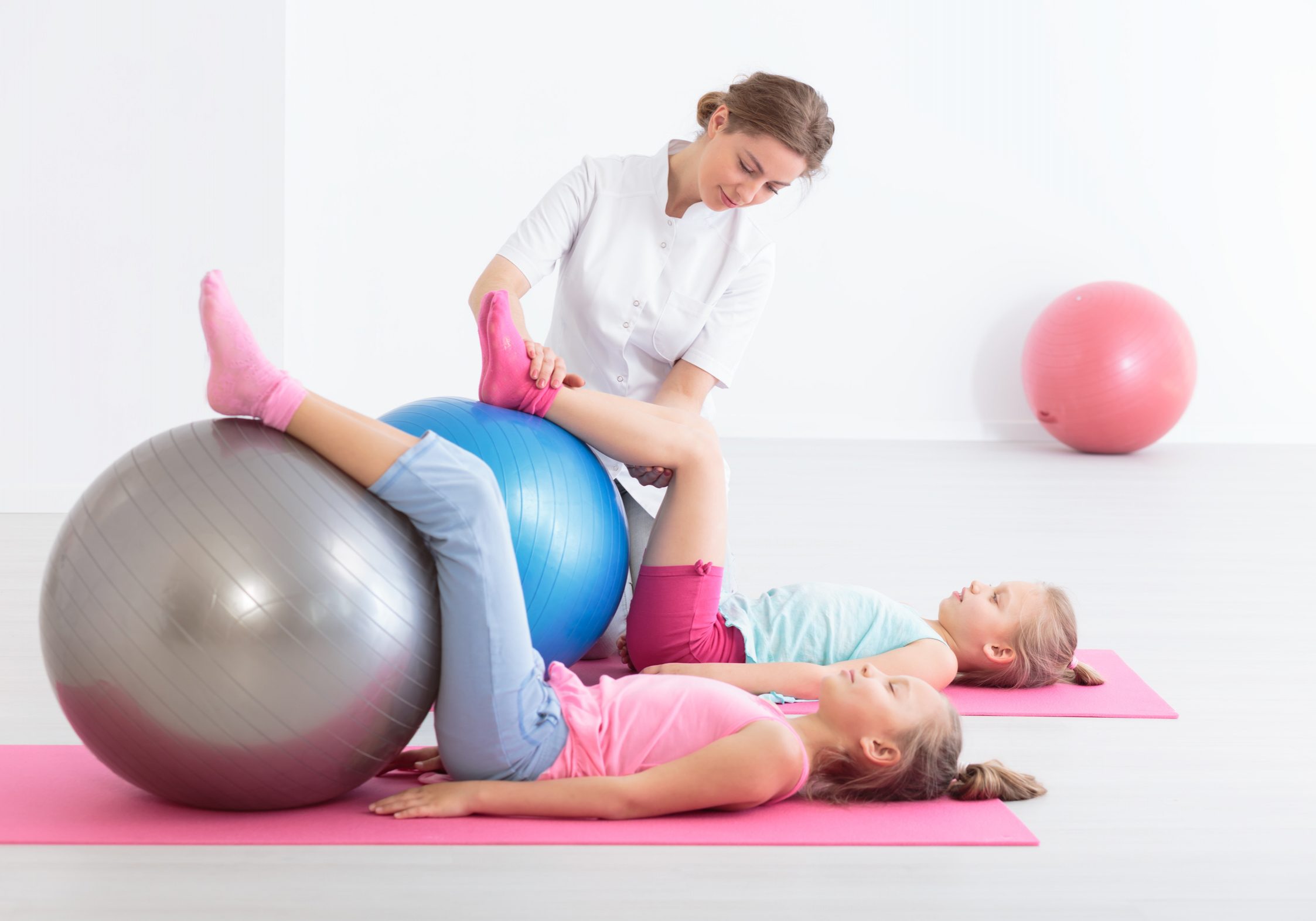 Two school girls lying on exercise mats exercising their legs on balls, accompanied by a physical therapist